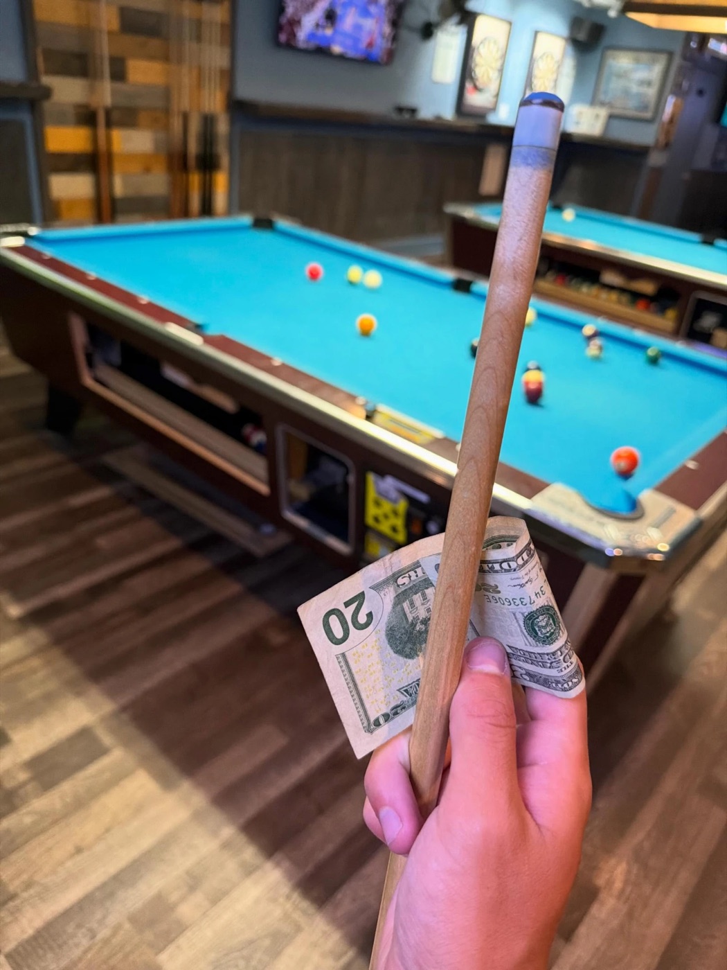 A hand holding a pool cue and a folded twenty-dollar bill in front of a pool table