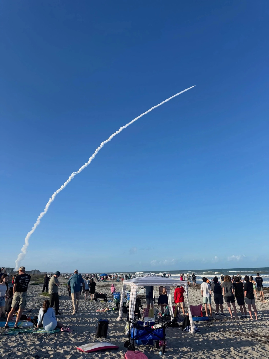 A rocket launch arcing a long smoke trail over a crowded Florida beach