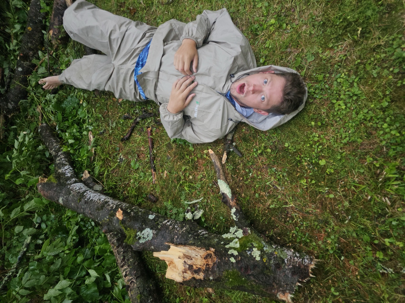 Kadin lying on the forest floor next to a fallen tree in a rain suit, mouth open in mock shock